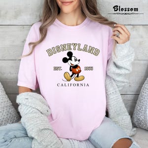 Puede incluir: Camiseta rosa con un gr&aacute;fico de estilo vintage de Mickey Mouse frente al logotipo de Disneyland. El texto dice "Disneyland Est. 1955 California".