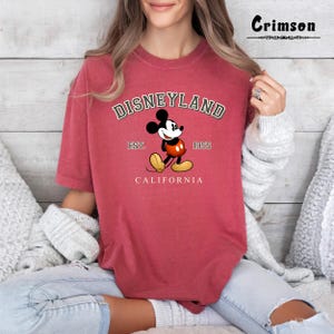 Puede incluir: Camiseta roja con un gr&aacute;fico de estilo vintage de Mickey Mouse frente al logotipo de Disneyland. El texto dice "DISNEYLAND EST. 1955 CALIFORNIA".