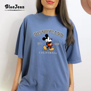 Puede incluir: Una camiseta azul claro con un gr&aacute;fico de estilo vintage de Mickey Mouse frente a un logotipo de Disneyland. El logotipo dice "DISNEYLAND EST. 1955 CALIFORNIA".