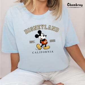 Puede incluir: Camiseta azul claro con un gr&aacute;fico de Mickey Mouse y el texto "DISNEYLAND EST. 1955 CALIFORNIA" impreso en la parte delantera.