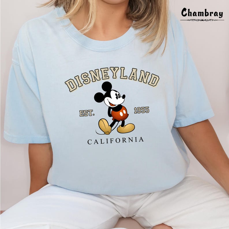 Puede incluir: Camiseta azul claro con un gr&aacute;fico de Mickey Mouse y el texto "DISNEYLAND EST. 1955 CALIFORNIA" impreso en la parte delantera.