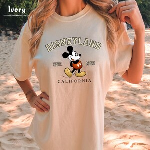 Puede incluir: Una camiseta beige con un gr&aacute;fico de Mickey Mouse y el texto "DISNEYLAND EST. 1955 CALIFORNIA".