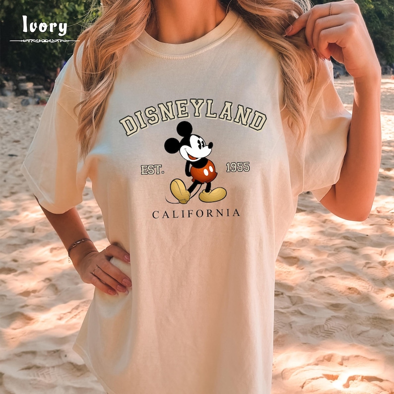 Puede incluir: Una camiseta beige con un gr&aacute;fico de Mickey Mouse y el texto "DISNEYLAND EST. 1955 CALIFORNIA".
