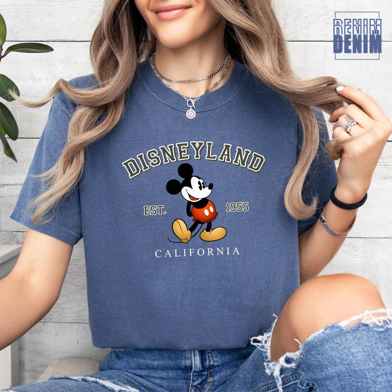 Puede incluir: Una camiseta azul con un gr&aacute;fico de estilo vintage de Mickey Mouse frente al logotipo de Disneyland. El texto dice "DISNEYLAND EST. 1955 CALIFORNIA".