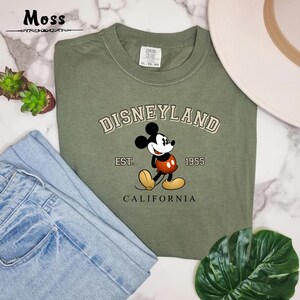 Puede incluir: Una camiseta verde con un logotipo vintage de Disneyland con Mickey Mouse. El logotipo dice "Disneyland Est. 1955 California".