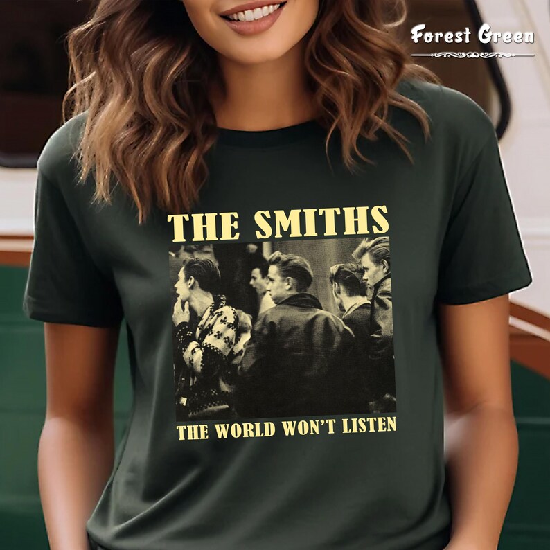 May include: A dark green t-shirt with a black and white image of The Smiths band members. The text "THE SMITHS" and "THE WORLD WON'T LISTEN" is printed in yellow on the shirt.
