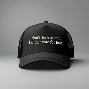 May include: Black trucker hat with a curved brim and mesh back. The hat features the white text, "don't look at me, I didn't vote for him" across the front. The hat is set against a gray background.