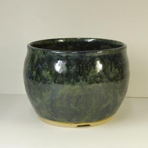 May include: A handmade ceramic bowl with a glossy, mottled glaze in shades of dark green and blue. The bowl has a rounded shape and a light-colored base. This decorative bowl is suitable for serving or display.