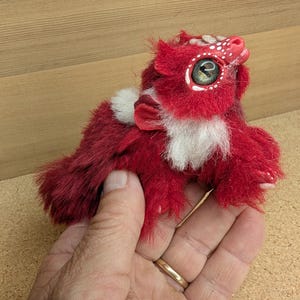 May include: A handmade plush creature with red fur, white accents, and a small red bow. The creature has large, detailed eyes and a spotted head. The plush is held in a hand, showcasing its size and craftsmanship.
