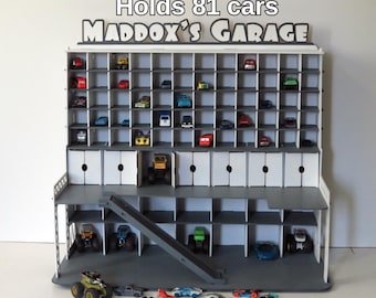 May include: A gray and white toy car garage with the text "Holds 81 cars" and "Maddox's Garage". The garage has multiple compartments to hold toy cars, with a ramp and doors. Several toy cars are visible inside the garage.