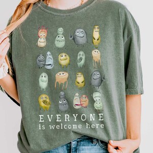 May include: Moss green unisex t-shirt with a watercolor illustration of various cartoon characters. The text "EVERYONE is welcome here" is printed below the characters. The shirt is made of a soft, comfortable material.