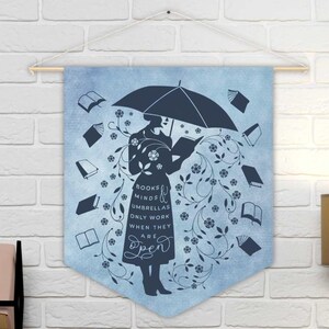 May include: Sky blue wall hanging with a silhouette of a person holding an umbrella, surrounded by books and floral designs. The text on the wall hanging reads "Books minds umbrellas only work when they are open."