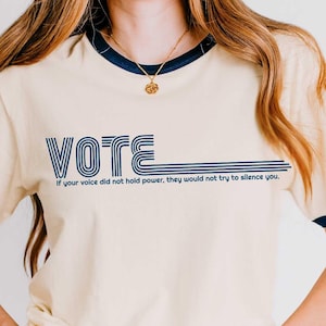 Op de afbeelding: Een naturelkleurig ringer-t-shirt met donkerblauwe bies op de kraag en mouwen. De voorkant toont het woord VOTE in een retro, meerlijnig lettertype, gevolgd door de tekst: If your voice did not hold power, they would not try to silence you.