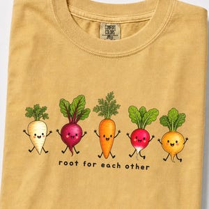 May include: A mustard-colored t-shirt featuring a graphic of five cartoon vegetables with arms and legs, including a parsnip, beet, carrot, radish, and turnip. Below the vegetables, the text reads "root for each other" in black letters.