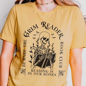 Könnte beinhalten: Senffarbenes T-Shirt mit einer Grafik, die ein Skelett beim Lesen eines Buches zeigt, umgeben von dem Text "Grim Reader Book Club" und "Beware the Reading is in our Bones". Das Shirt hat ein Unisex-Größenetikett.