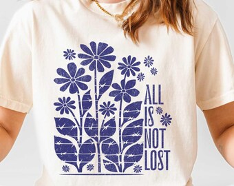 May include: Ivory-colored unisex t-shirt with a blue floral design and the text "ALL IS NOT LOST." The shirt is made of a soft material and has a relaxed fit. The model is wearing light blue jeans and white sneakers.