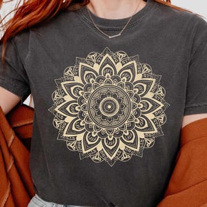 May include: Dark gray unisex t-shirt featuring a beige mandala design. The shirt has short sleeves and a crew neck. The image also shows a brown jacket and blue jeans. The text "UNISEX SIZING" is visible at the top.
