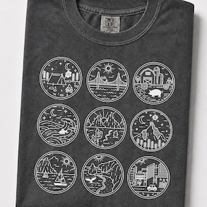 Könnte beinhalten: Ein graphitgraues T-Shirt mit einer weißen Linienzeichnung, die neun kreisförmige Szenen zeigt. Die Motive umfassen einen Campingplatz, eine Brücke, einen Bauernhof mit Schwein, Meereswellen, eine Wüstenlandschaft, einen Berg, ein Segelboot, eine Küste und eine Stadt.
