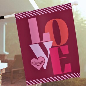 Puede incluir: Una bandera de jardín de color rojo baya con la palabra "LOVE" en tonos rosas y rojos. La letra "V" tiene un corazón con la palabra "always". La bandera tiene un borde a rayas y se muestra al aire libre.