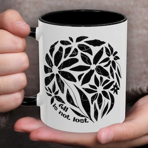 May include: White ceramic mug with a black rim and handle. The mug features a black and white graphic of stylized flowers with the text "All is not lost."