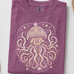 May include: A berry-colored t-shirt featuring a cream-colored graphic of a jellyfish with intricate patterns. The text reads: If injustice becomes law resistance becomes duty, defend democracy, speak truth to power, and love trumps hate.