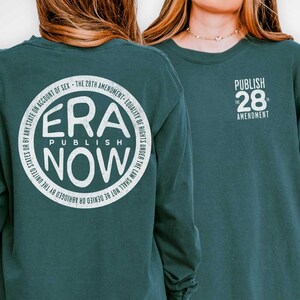 Può includere: Maglietta a maniche lunghe blu abete con testo bianco. La parte anteriore della maglietta recita "PUBLISH 28 AMENDMENT". La parte posteriore della maglietta presenta un design circolare con "ERA PUBLISH NOW" e testo sul bordo. La maglietta è realizzata in materiale Comfort Colors.