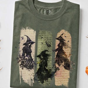 May include: An olive green crew neck t-shirt featuring a graphic print of three witches in pointed hats. The design includes the text 1692 and layered vintage-style document text in the background. The shirt has a ribbed collar and short sleeves.