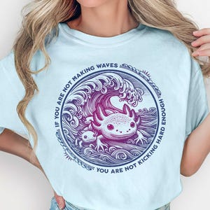 May include: Pale blue t-shirt with a circular graphic of an axolotl in a wave. The text reads, "If you are not making waves, you are not kicking hard enough." The shirt is labelled "Unisex Sizing."