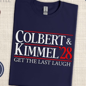 May include: Navy blue t-shirt with the text "COLBERT & KIMMEL '28 GET THE LAST LAUGH" in white and red. The shirt is next to a pair of sunglasses, a "VOTE" button, and a "NAVY" badge. The shirt is labeled "unisex sizing".