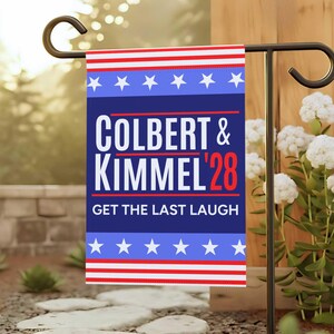 Può includere: Una bandiera da giardino, 30 x 46 cm, presenta il testo "COLBERT & KIMMEL '28 GET THE LAST LAUGH" in bianco su sfondo blu, con strisce e stelle rosse e bianche. La bandiera è su un supporto in metallo nero.