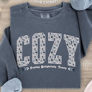 May include: A denim-colored sweatshirt with the word "COZY" in a floral pattern across the front. The text "Feeling Delightfully Toasty" is below the word "COZY". The sweatshirt has rolled-up sleeves and the text "denim" is at the bottom.