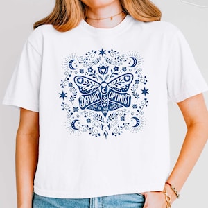 May include: A white boxy cropped t-shirt featuring a dark blue graphic print of a moth surrounded by stars, moons, and floral patterns. The text on the moth reads "DEFIANT OPTIMIST" and "all is not lost" in a stylized, decorative font.