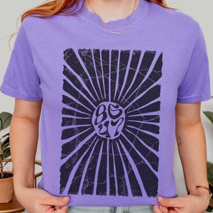 Könnte beinhalten: Ein violettes Comfort Colors T-Shirt mit einem schwarzen Sonnenstrahl-Design. Der Sonnenstrahl hat eine Used-Optik und ein zentrales Design mit dem Text "86 27". Der Text "more colors!" ist sichtbar. Das Wort "violet" ist auf der linken Seite.