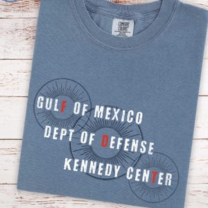 Puede incluir: Una camiseta azul vaquero con el texto "GULF OF MEXICO DEPT OF DEFENSE KENNEDY CENTER" en blanco y rojo. La camiseta tiene una etiqueta de tallas unisex y una etiqueta "BLUE JEAN".