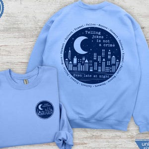 Può includere: Due felpe unisex blu Carolina. Una presenta una grafica circolare con una luna crescente, uno skyline e il testo "Telling Jokes is not a crime, Even late at night." L'altra ha una versione più piccola della stessa grafica.
