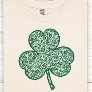 May include: Cream-coloured t-shirt with a large green shamrock design. The shamrock is filled with detailed floral patterns. The shirt has a crew neck and short sleeves. The text "UNISEX SIZING" is at the top left.