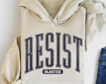 Pode incluir: Moletom unissex cor areia com a palavra "RESIST" em letras de bloco azul marinho. Abaixo está o texto "INJUSTICE". Uma calça jeans azul é visível no lado direito do moletom dobrado.