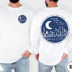 May include: White long-sleeved shirt featuring a navy blue graphic on the front and back. The front graphic is a circular design with a crescent moon and cityscape. The back graphic says "Telling Jokes is not a crime" with a moon and cityscape.