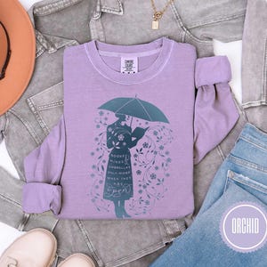 May include: Orchid-colored sweatshirt featuring a teal illustration of a person holding an umbrella and reading a book. The text on the shirt reads "Books Minds Umbrellas Only Work When They Are Open". The sweatshirt is displayed with a hat, jacket, and jeans.