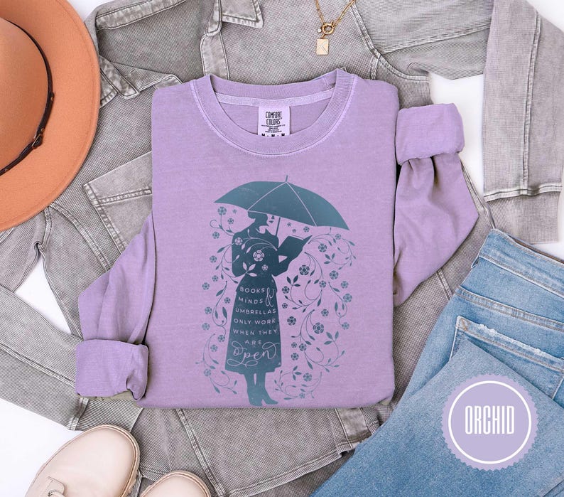 May include: Orchid-colored sweatshirt featuring a teal illustration of a person holding an umbrella and reading a book. The text on the shirt reads "Books Minds Umbrellas Only Work When They Are Open". The sweatshirt is displayed with a hat, jacket, and jeans.