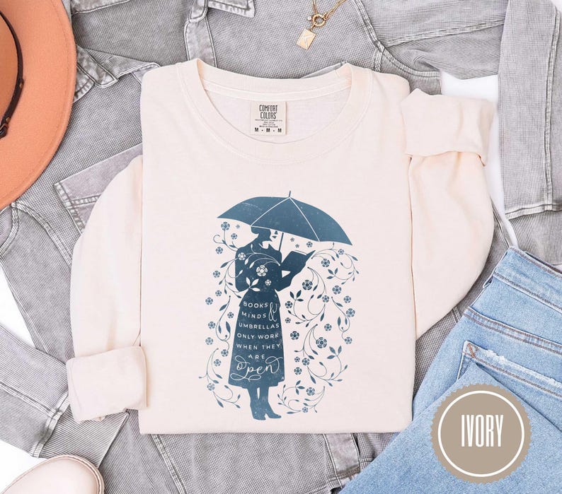 May include: Ivory-colored long-sleeve sweatshirt with a teal graphic of a person holding an umbrella and reading a book. The text on the shirt reads "Books, Minds, Umbrellas Only Work When They Are Open."