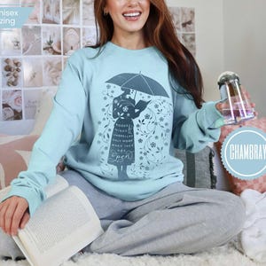 May include: A light blue long-sleeve shirt with a graphic of a person holding an umbrella and reading a book. The text on the shirt reads "Books & Minds & Umbrellas Only Work When They Are Open". The shirt is labeled "unisex sizing".