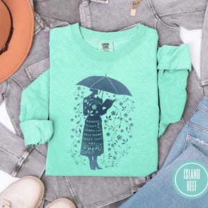 May include: A mint green long-sleeve shirt with a graphic of a person under an umbrella reading a book. The graphic includes the text "BOOKS MINDS UMBRELLAS ONLY WORK WHEN THEY ARE OPEN". The shirt is from Island Reef.