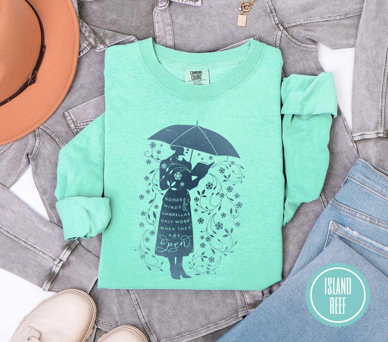 May include: A mint green long-sleeve shirt with a graphic of a person under an umbrella reading a book. The graphic includes the text "BOOKS MINDS UMBRELLAS ONLY WORK WHEN THEY ARE OPEN". The shirt is from Island Reef.