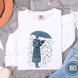 May include: White long-sleeve sweatshirt with a teal graphic of a person under an umbrella reading a book. The graphic includes the text "Books, minds, umbrellas only work when they are open." A white circle with the word "WHITE" is in the bottom right.