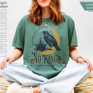 Woman wearing light green (medium muted green) Comfort Colors tees featuring a dark blue graphic of a raven, crown, and moon with No Kings – History is Watching design; Graphic is dark blue with goldenrod accents and crescent moon. unisex sizing displayed.