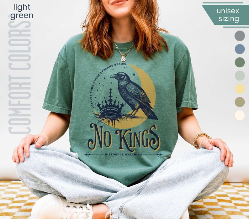 Woman wearing light green (medium muted green) Comfort Colors tees featuring a dark blue graphic of a raven, crown, and moon with No Kings – History is Watching design; Graphic is dark blue with goldenrod accents and crescent moon. unisex sizing displayed.