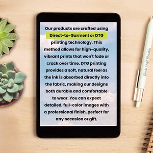 May include: A tablet displays text about Direct-to-Garment (DTG) printing technology, highlighting high-quality, vibrant prints. A white pen with the text "Quate Craft Creative" and a spiral notebook are on a wooden surface, along with two potted succulents.