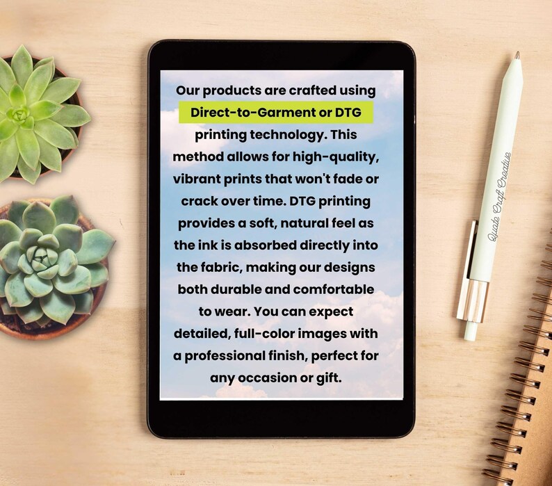 May include: A tablet displays text about Direct-to-Garment (DTG) printing technology, highlighting high-quality, vibrant prints. A white pen with the text "Quate Craft Creative" and a spiral notebook are on a wooden surface, along with two potted succulents.