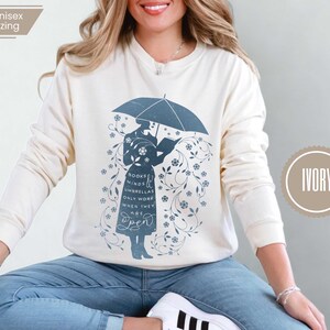 May include: Ivory-colored sweatshirt featuring a blue illustration of a person under an umbrella reading a book. The design includes floral accents and the text "Books Minds Umbrellas Only Work When They Are Open."